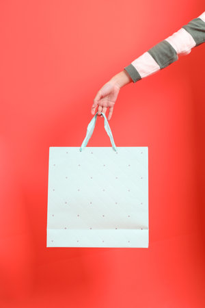 A close-up of a hand wearing a green stripe pattern sweater holding up a shopping bag, isolated against a red background. Merry Christmas.の写真素材