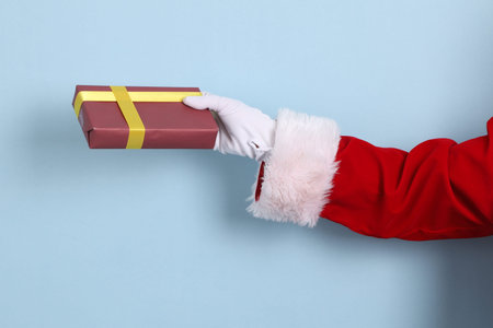 A close-up of a hand of Santa Claus holding up a gift or present box, isolated against a blue background. Merry Christmas.の写真素材