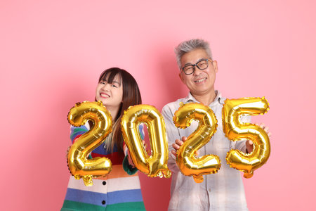 Asian senior man and young woman in casual look hand holding 3d golden metallic balloons forming the number 2025 with two thousand twenty-five New Year isolated on pink background.の写真素材
