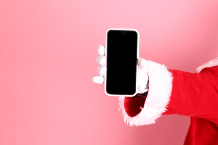 Merry Christmas, hand of senior man celebrate winter holidays is holding mobile phone, wearing Santa Claus costume isolated on pink background. Merry Xmasの写真素材