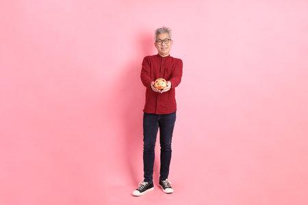 Happy Chinese New Year, asian senior man in formal and casual red shirts with gesture of  holding Chinese piggy bank isolated on the pink background.の写真素材