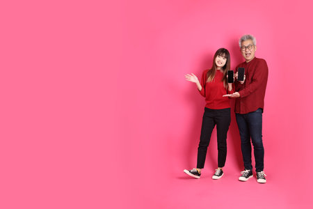 Chinese New Year, Asian senior man and woman wear santa hat, casual red clothes with gesture of Holding mobile phone, pointing isolated on pink background.の写真素材