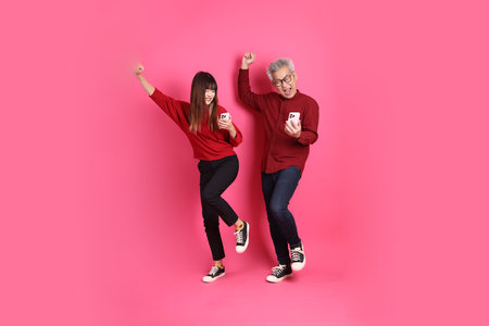 Chinese New Year, Asian senior man and woman wear santa hat, casual red clothes with gesture of Holding mobile phone, yes, excited, celebrating isolated on pink background.の写真素材