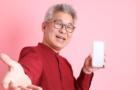 Happy Chinese New Year, asian senior man in formal and casual red shirts with gesture of Holding mobile phone, taking a selfie or video call isolated on pink background.の写真素材