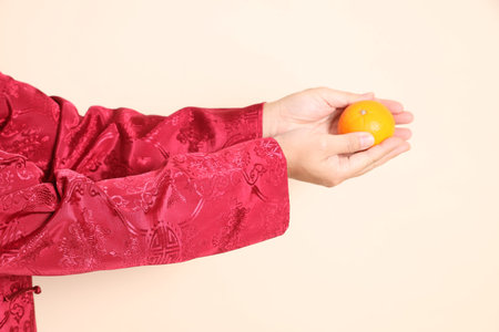 Asian Chinese energetic senior man wearing golden traditional cheongsam qipao or changshan dress with gesture of hand holding Tangerine or Orange isolated on beige background.の写真素材
