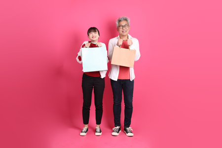 St Valentine's Day, Asian senior and young woman wear casual red clothes with white heart-shaped shirt jacket with gesture of holding shopping bag isolated on pink background.の写真素材