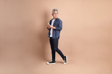 Asian senior man in casual clothes with gesture of Holding mobile phone isolated on brown background,. Smart casual.の写真素材