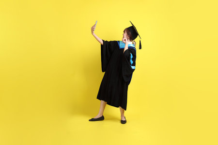 Female university student in graduation gowns and caps with gesture of taking a selfie isolated on yellow background.の写真素材
