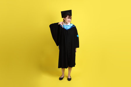 Female university student in blue graduation gowns and caps isolated on yellow background. Gen Z, fresh graduate, education, first jobberの写真素材
