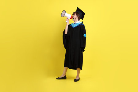 Female university student in blue graduation gowns and caps with gesture of shouting, holding megaphone isolated on yellow background.の写真素材