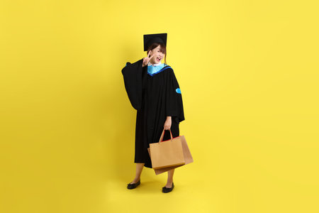Female university student in graduation gown and cap with gesture of holding shopping bag isolated on yellow background. Gen Z, fresh graduate, education, first jobberの写真素材