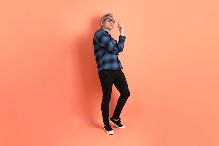 Asian senior man in Green plaid casual clothes wearing glasses with gesture of Standing and smiling isolated on orange background.の写真素材