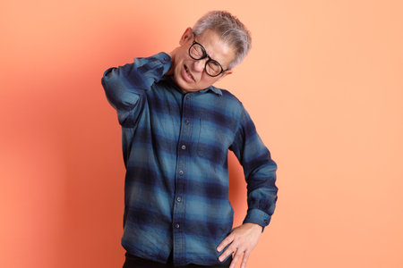Asian senior man in plaid casual clothes wearing glasses with gesture of backache isolated on orange background.の写真素材