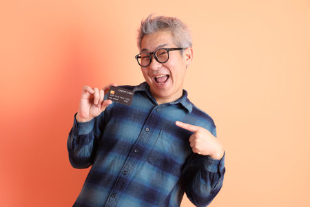 Asian senior man in green plaid casual clothes wearing glasses with gesture of holding credit card isolated on orange background. Smart casual.の写真素材