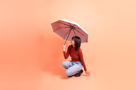 Short hair asian woman in red casual long sleeve t-shirt with gesture of hand Holding blue and pink Umbrella isolated on orange background. Gen Zの写真素材