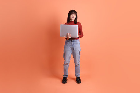 Short hair asian woman in red casual long sleeve t-shirt with gesture of Holding laptop isolated on orange background. Gen Zの写真素材