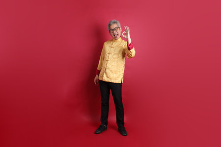 Asian Chinese senior man wearing golden traditional cheongsam qipao or changshan dress with gesture of thumbs up, ok or confirm isolated on red background.の写真素材