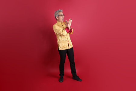 Asian Chinese senior man wearing golden traditional cheongsam qipao or changshan dress with gesture of stop, isolated on red background.の写真素材