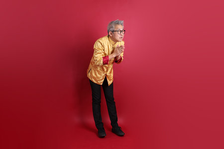Asian Chinese senior man wearing golden traditional cheongsam qipao or changshan dress isolated on red background.の写真素材