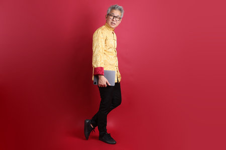 Happy Chinese new year. Asian Chinese senior man wearing golden traditional cheongsam qipao or changshan dress with gesture of holding tablet isolated on red background.の写真素材