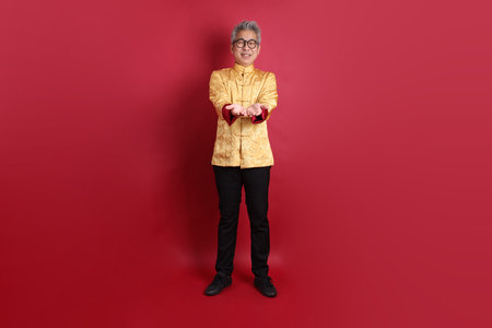 Happy Chinese new year. Asian Chinese senior man wearing golden traditional cheongsam qipao or changshan dress with gesture of giving and receiving isolated on red background.の写真素材