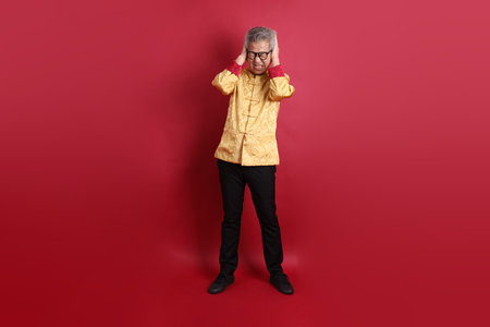 Asian Chinese senior man wearing golden traditional cheongsam qipao or changshan dress with gesture of ear close isolated on red background.の写真素材