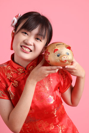 Asian Chinese woman wearing red traditional cheongsam qipao with gesture of holding gold piggy bank isolated on pink background.の写真素材