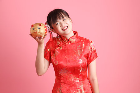 Happy Chinese new year. Asian Chinese woman wearing red traditional cheongsam qipao with gesture of holding gold Chinese piggy bank isolated on pink background. Gen Zの写真素材