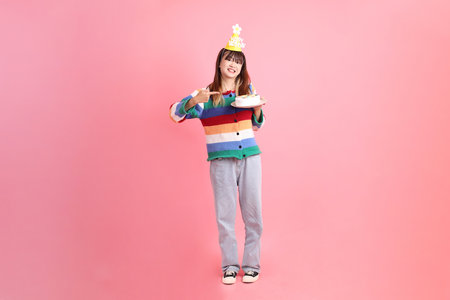 HAPPY BIRTHDAY, Asian woman with colorful rainbow winter clothes , casual look, birthday hat with gesture of Holding birthday cake isolated on pink background. Gen Z,の写真素材