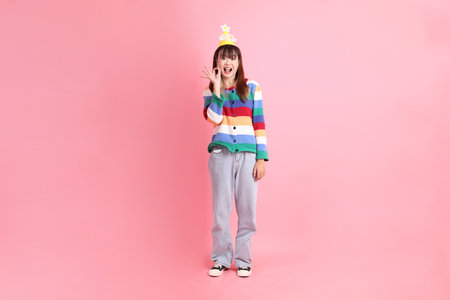 Asian woman with colorful rainbow winter clothes, casual look, birthday hat with gesture of thumbs up, ok or confirm isolated on pink background. Gen Z,の写真素材