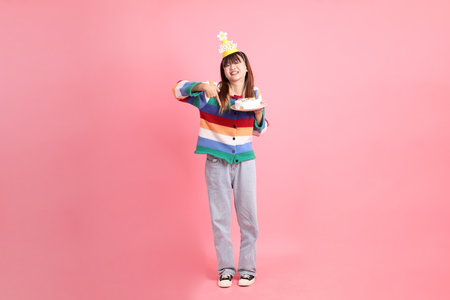 Asian woman with colorful rainbow winter clothes, casual look, birthday hat with gesture of holding birthday cake isolated on pink background. Gen Z,の写真素材