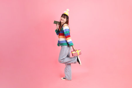 HAPPY BIRTHDAY, Asian woman with colorful rainbow winter clothes , casual look, birthday hat with gesture of Holding gift box isolated on pink background. Gen Zの写真素材
