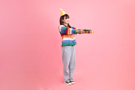 HAPPY BIRTHDAY, Asian woman with colorful rainbow winter clothes , casual look, birthday hat with gesture of Holding gift box isolated on pink background. Gen Zの写真素材