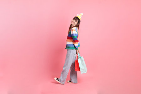 HAPPY BIRTHDAY, Asian woman with colorful rainbow winter clothes , casual look, birthday hat with gesture of holding shopping bag isolated on pink background. Gen Z,の写真素材