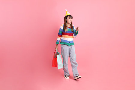 Asian woman with colorful rainbow winter clothes, casual look, birthday hat with gesture of holding shopping bag isolated on pink background. Gen Z,の写真素材