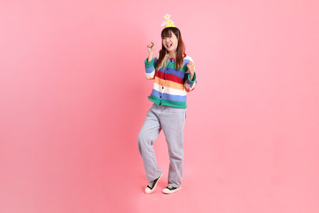 HAPPY BIRTHDAY, Asian woman with colorful rainbow winter clothes , casual look, birthday hat with gesture of yes, excited, celebrating isolated on pink background. Gen Z,の写真素材