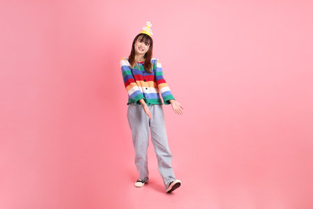 HAPPY BIRTHDAY, Asian woman with colorful rainbow winter clothes , casual look, birthday hat with gesture of pointing isolated on pink background. Gen Z,の写真素材