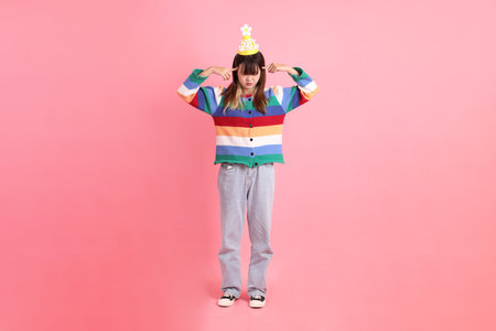 HAPPY BIRTHDAY, Asian woman with colorful rainbow winter clothes , casual look, birthday hat with gesture of thinking isolated on pink background. Gen Z,の写真素材