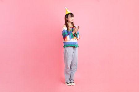 HAPPY BIRTHDAY, Asian woman with colorful rainbow winter clothes , casual look, birthday hat with gesture of claping hands isolated on pink background. Gen Z,の写真素材