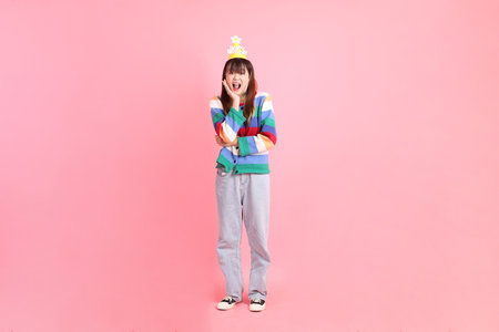 HAPPY BIRTHDAY, Asian woman with colorful rainbow winter clothes , casual look, birthday hat with gesture of surprise, Wow and impressive isolated on pink background. Gen Z,の写真素材