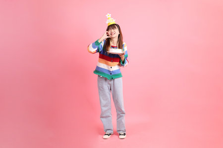 HAPPY BIRTHDAY, Asian woman with colorful rainbow winter clothes , casual look, birthday hat with gesture of Holding birthday cake isolated on pink background. Gen Z,の写真素材