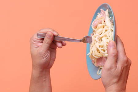 Hand holding fork eating creamy spaghetti carbonara on a green plate, isolated on an orange background. Italian food, Italian cuisine, noodles.の写真素材