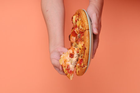 Hand holding a whole Hawaiian pizza, a pizza topped with tomato sauce, cheese, pineapple, and ham or bacon on a white plate, isolated on an orange background.の写真素材