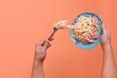 Hand holding fork eating creamy spaghetti carbonara on a green plate, isolated on an orange background. Italian food, Italian cuisine, noodles.の写真素材
