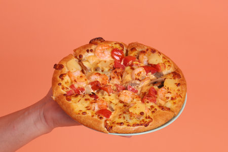 Hand holding a whole Hawaiian pizza, a pizza topped with tomato sauce, cheese, pineapple, and ham or bacon on a white plate, isolated on an orange background.の写真素材