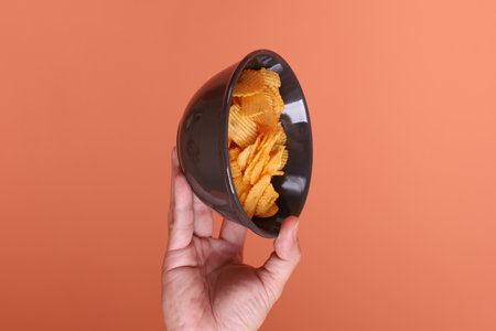Hand holding potato chips on a black bowl, isolated on an orange background. junk food, snack, studio.の写真素材