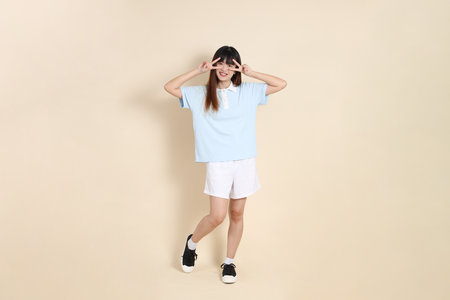 Full body portrait of a young Asian woman wearing a light blue polo shirt, and white shorts, peace sign, V Sign, two finger against a plain beige background. College student, Gen Zの写真素材