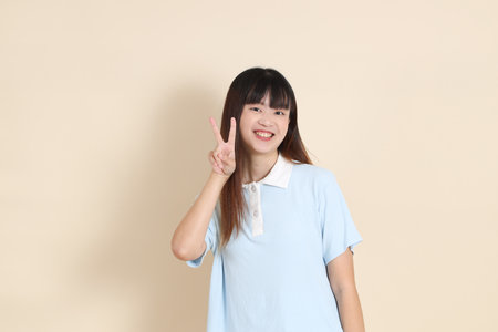 Medium shot of a young Asian woman wearing a light blue polo shirt, peace sign, V Sign, two finger against a plain beige background. College student, Gen Zの写真素材