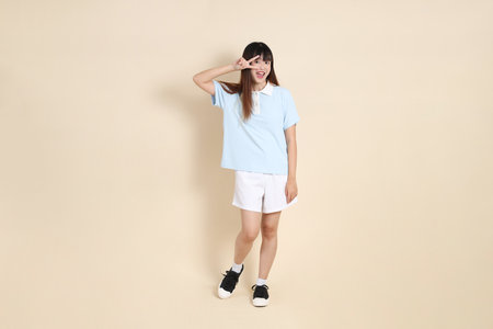 Full body portrait of a young Asian woman wearing a light blue polo shirt, and white shorts, peace sign, V Sign, two finger against a plain beige background. College student, Gen Zの写真素材