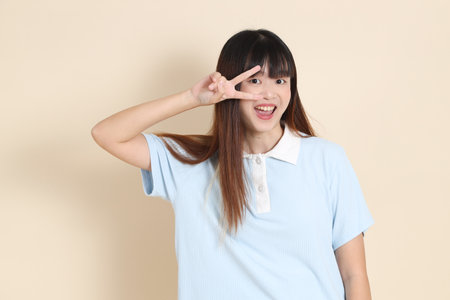 Medium shot of a young Asian woman wearing a light blue polo shirt, peace sign, V Sign, two finger against a plain beige background. College student, Gen Zの写真素材